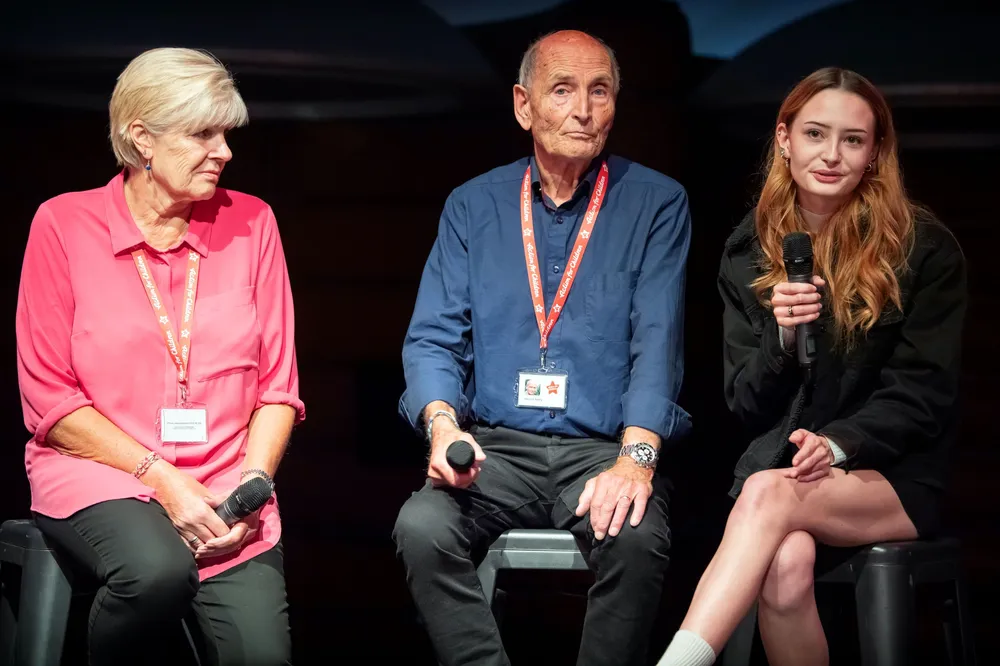 Three people on stools speaking in front of a black background from left to right: an older woman, an older man, and a younger woman with a microphone in her hand