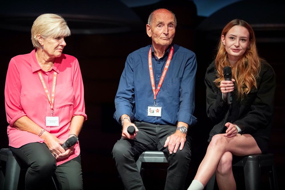 Three people on stools speaking in front of a black background from left to right: an older woman, an older man, and a younger woman with a microphone in her hand