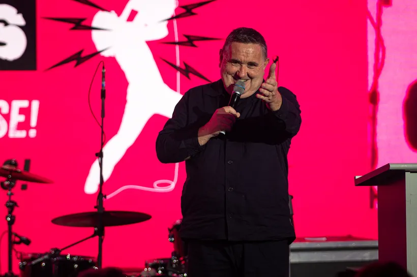 man with black shirt on stage with a mic in his hand and hand infront of him