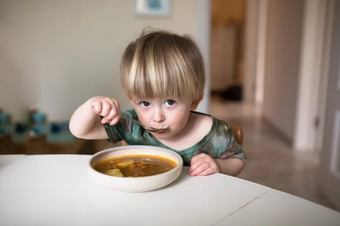 Young boy looking at camera whilst sitting at a white table eating a spoonful of food from a bowl