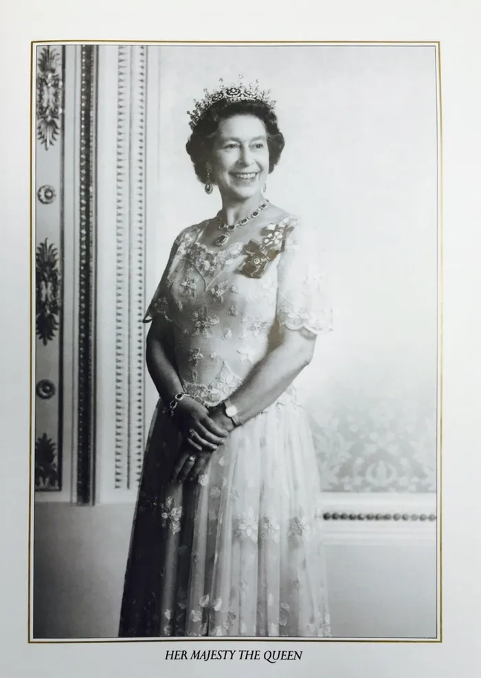 Black and white formal portrait of Queen Elizabeth II wearing an ornate tiara and an elegant lace gown adorned with floral patterns and medals. The setting includes a decorated wall with intricate molding, emphasizing a regal and dignified atmosphere.