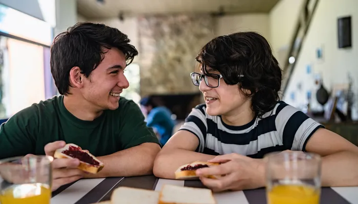 Two boys sitting at a table, each holding a slice of bread with jam, engaged in conversation. The table has additional slices of bread and glasses of orange juice, with a cozy indoor setting featuring a stone fireplace in the blurred background.