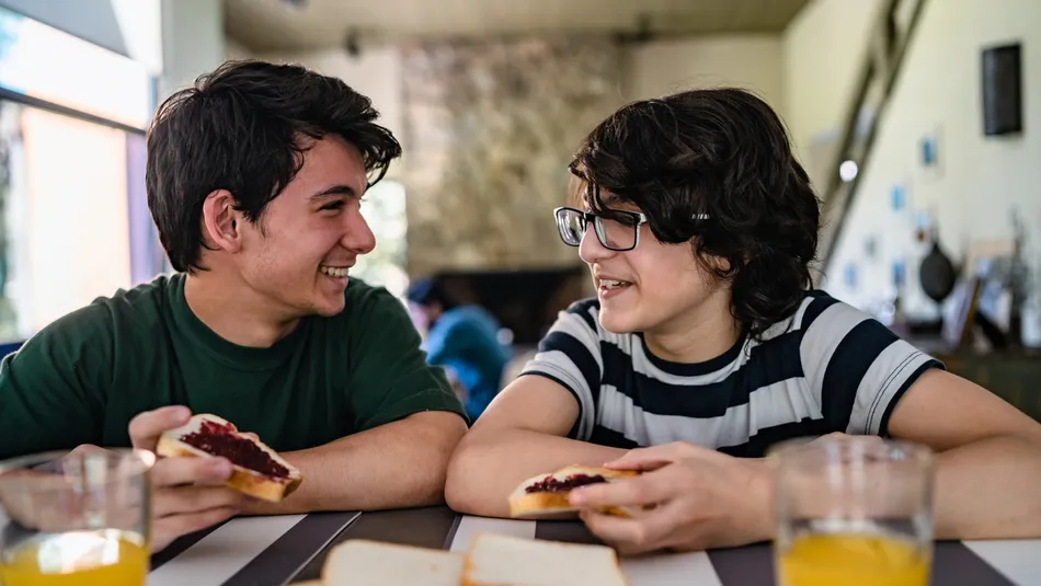 Two boys sitting at a table, each holding a slice of bread with jam, engaged in conversation. The table has additional slices of bread and glasses of orange juice, with a cozy indoor setting featuring a stone fireplace in the blurred background.