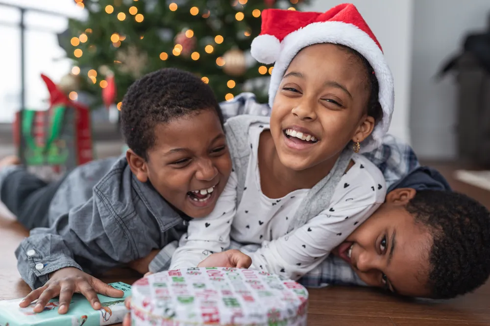 Three siblings laughing and laying on top of each other with Christmas gift presents in front of them a lit up tree in the background