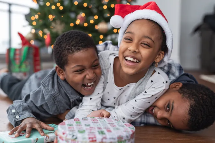 Three siblings laughing and laying on top of each other with Christmas gift presents in front of them a lit up tree in the background