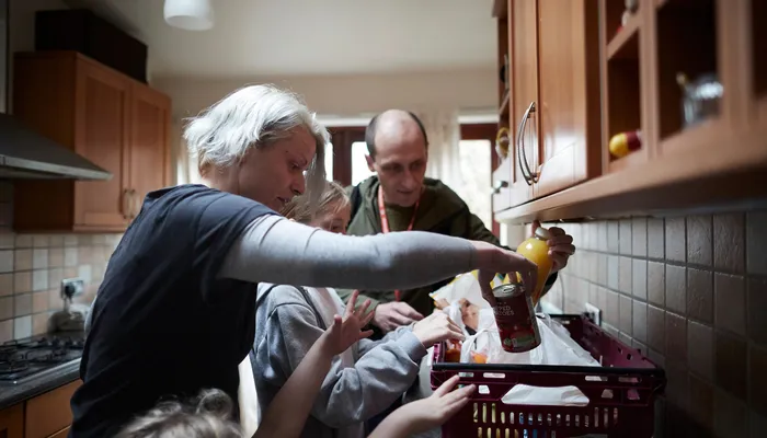 Two adults and two children in a kitchen unpacking groceries from a plastic crate on the worktop. One of the adults is wearing a red lanyard with a blurred logo, possibly suggesting a food donation from a charitable organisation.
