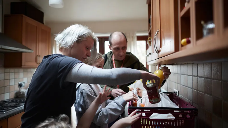 Two adults and two children in a kitchen unpacking groceries from a plastic crate on the worktop. One of the adults is wearing a red lanyard with a blurred logo, possibly suggesting a food donation from a charitable organisation.