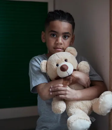 A child wearing a gray t-shirt hugging a light brown teddy bear. Background includes green shuttered doors on the left and wooden panelling on the right, suggesting an indoor setting.