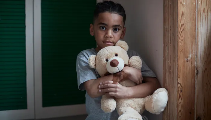 A child wearing a gray t-shirt hugging a light brown teddy bear. Background includes green shuttered doors on the left and wooden panelling on the right, suggesting an indoor setting.