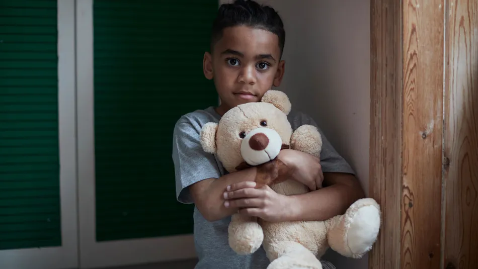 A child wearing a gray t-shirt hugging a light brown teddy bear. Background includes green shuttered doors on the left and wooden panelling on the right, suggesting an indoor setting.