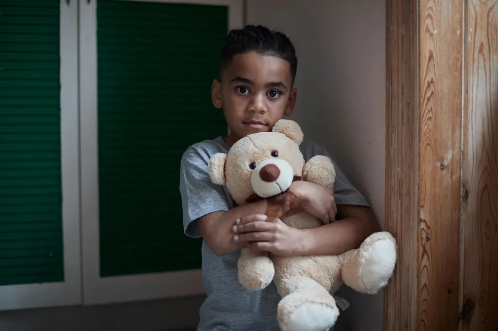 A child wearing a gray t-shirt hugging a light brown teddy bear. Background includes green shuttered doors on the left and wooden panelling on the right, suggesting an indoor setting.