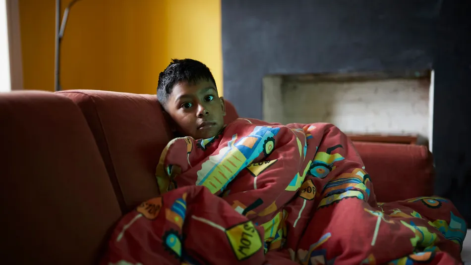 A young boy sitting on a red sofa wrapped in a colourful duvet with vehicle designs, set against a yellow wall and a dark fireplace background in a sparsely decorated room.