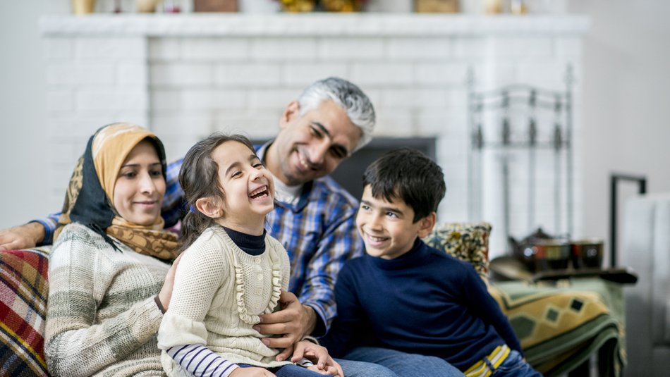 A Muslim mother, father, son and daughter are indoors in a living room. The parents are sitting on the sofa, and the daughter is laughing while being tickled.