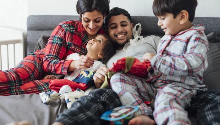 A family in matching checked pyjamas sitting closely on a bed, with a child lying on an adult's lap and another child holding a wrapped gift. The cozy setting suggests a relaxed, intimate moment, possibly during a holiday or special occasion.