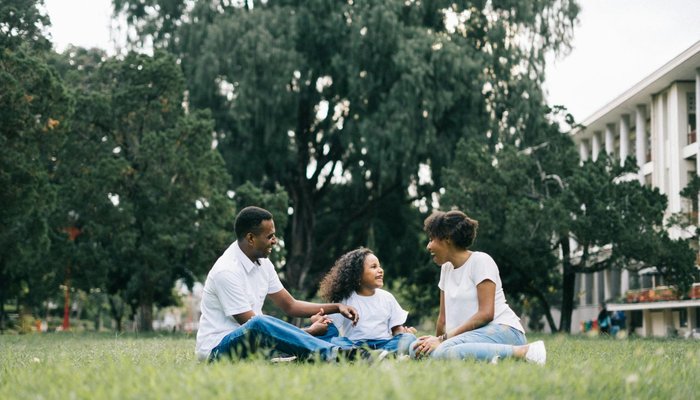 A family sitting on grass in a park-like setting with large trees and a building in the background. The family members are engaged in conversation, wearing casual white tops and blue jeans, conveying a relaxed and happy outdoor moment.