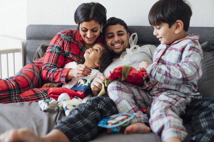 A family sitting on their bed during the Christmas period. The two young sons open presents as their parents watch smiling