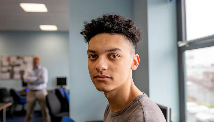 A young person with curly hair wearing a gray t-shirt, standing in a classroom setting featuring desks, chairs, and computer monitors near a large window with natural light. In the blurred background a man in casual business attire stands with arms folded
