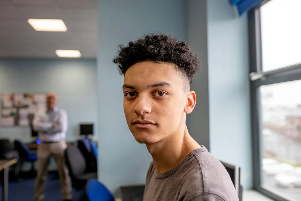 A young person with curly hair wearing a gray t-shirt, standing in a classroom setting featuring desks, chairs, and computer monitors near a large window with natural light. In the blurred background a man in casual business attire stands with arms folded