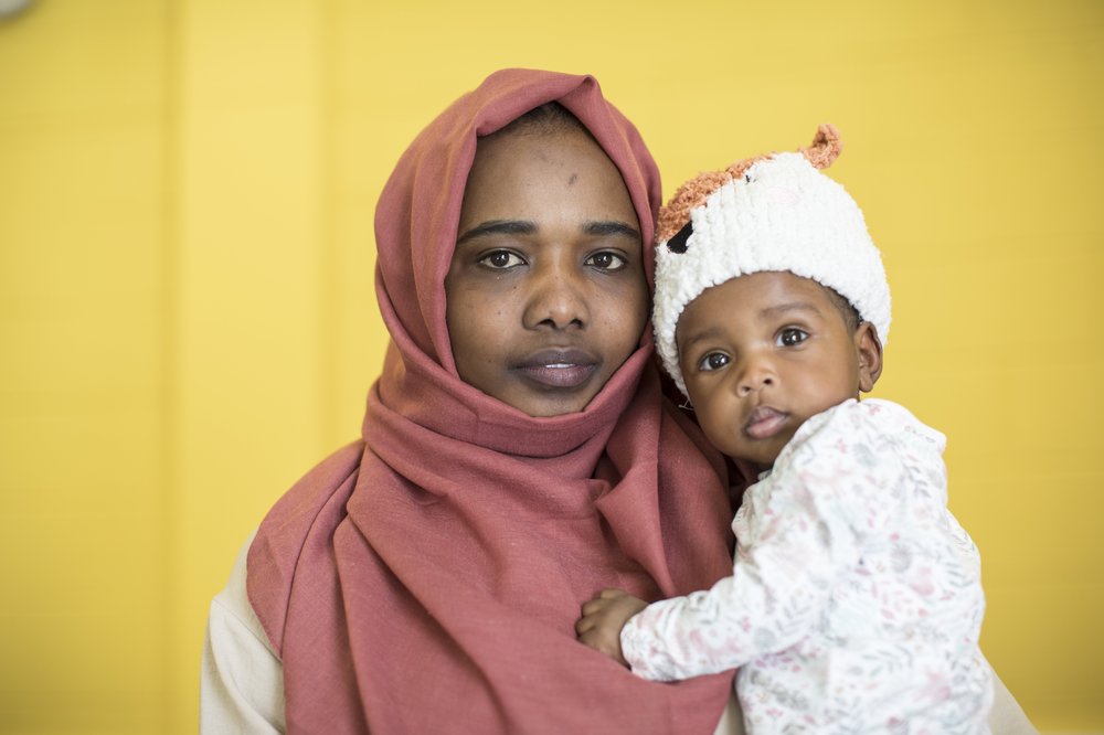 Samira, in a red head scarf, looks into the camera holding her baby daighter close to