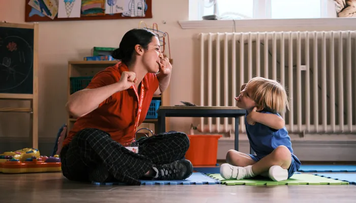A woman and a toddler sitting on foam mats in a playroom, engaging in a playful interaction with the woman making a gesture and the child mimicking or responding. The room features children's artwork on the wall, a radiator, and various toys