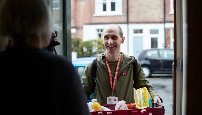 A person wearing an Action for Children lanyard holding a red basket filled with groceries, standing outside a doorway. The scene captures a delivery or assistance interaction, with a residential street and parked cars visible in the background