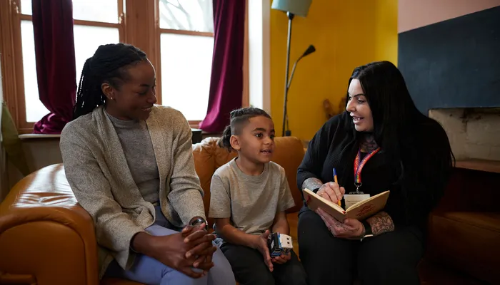 Two adults and a child sitting on a sofa. One person, wearing an Action for Children lanyard is holding a notebook and pen, appearing to take notes while engaging with the child and another adult, suggesting a meeting or counselling session.