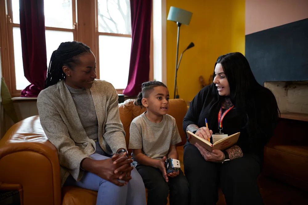 Two adults and a child sitting on a sofa. One person, wearing an Action for Children lanyard is holding a notebook and pen, appearing to take notes while engaging with the child and another adult, suggesting a meeting or counselling session.