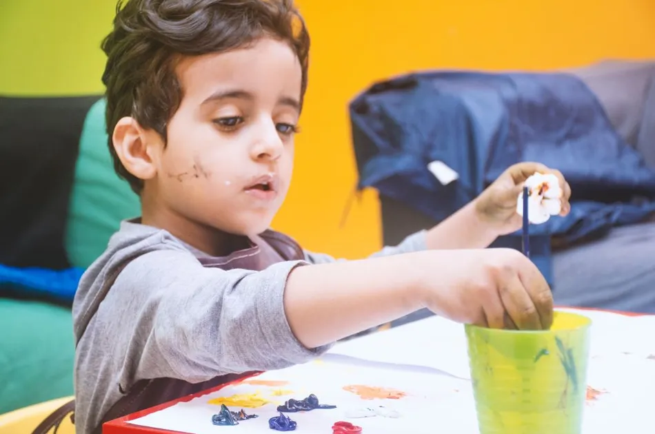 Young boy wearing an apron sitting at a table dipping a paint brush in a cup of water