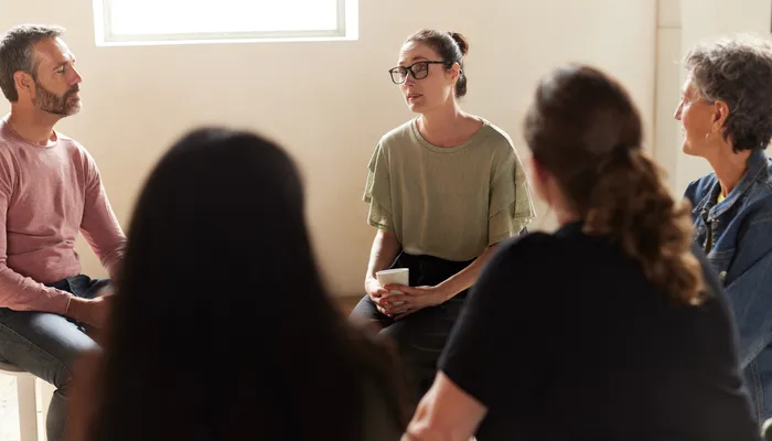 A group therapy session with five people sitting in a circle in a well-lit room, engaging in conversation. Participants display attentive postures, with one person holding a cup, indicating a supportive and interactive environment.