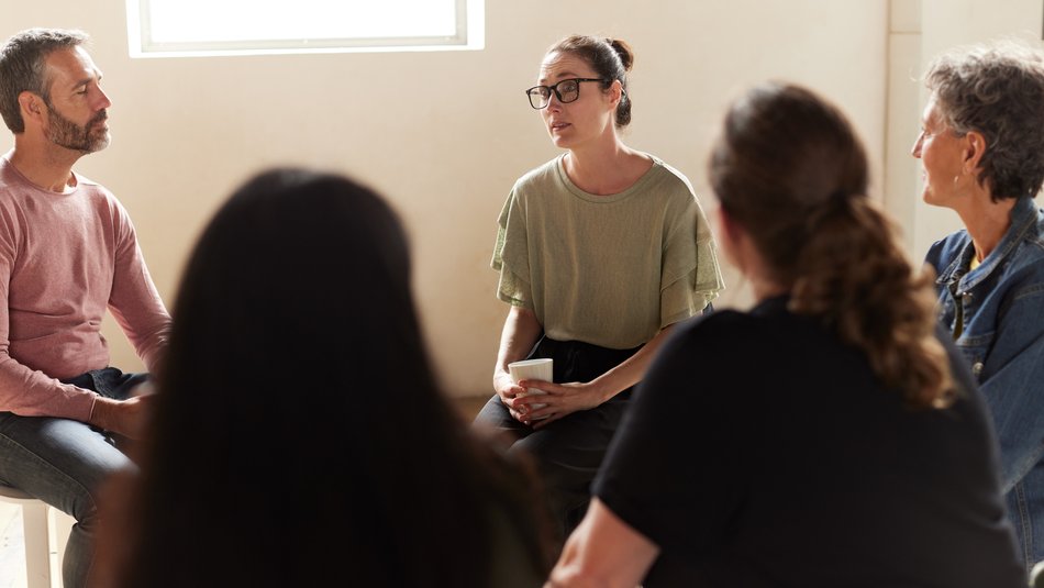 A group therapy session with five people sitting in a circle in a well-lit room, engaging in conversation. Participants display attentive postures, with one person holding a cup, indicating a supportive and interactive environment.