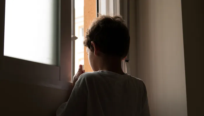 A young boy standing indoors, looking out through a partially open window with soft natural light illuminating his silhouette. The scene conveys a quiet, contemplative moment with muted colours and a focus on the boy’s back and profile.