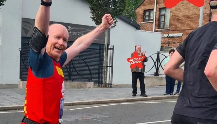 A street scene during a running event where a participant in a red vest raises both arms in celebration. A person in the background holds a red star-shaped sign with "Action for Children" text, indicating support for a children's charity.