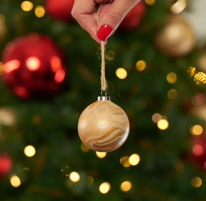 A hand with red nail polish holding a gold marbled Christmas ornament by a string in front of a decorated Christmas tree. Background features blurred red and gold ornaments and warm yellow lights, creating a festive holiday atmosphere.