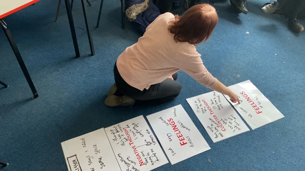 A women with her back to the camera kneeling on a workshop floor pointing at one of four A3 pieces of paper with writing on