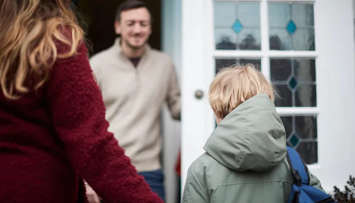 A child wearing a green jacket and backpack standing outside a house with a woman in a red sweater nearby and a man in a light-colored sweater at the doorway. The scene suggests a moment of arrival or departure, possibly related to school or a visit.