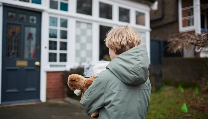 A child wearing a green jacket holding a teddy bear and a plastic bag while standing outside a house with a dark blue door and white-framed windows. The scene suggests the child is about to enter or has just arrived at the house