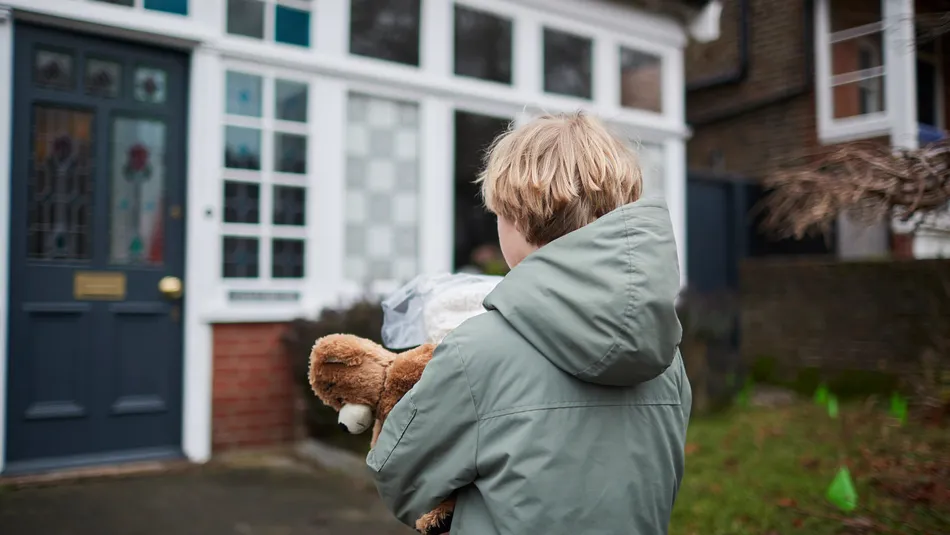 A child wearing a green jacket holding a teddy bear and a plastic bag while standing outside a house with a dark blue door and white-framed windows. The scene suggests the child is about to enter or has just arrived at the house