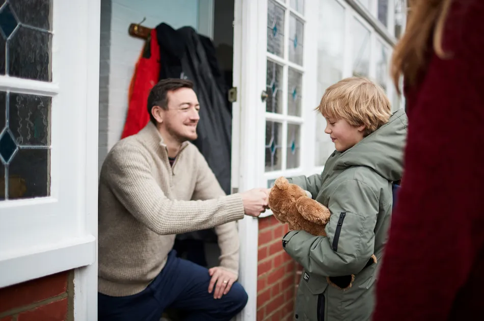 An adult kneeling at a doorway, handing an object to a child holding a teddy bear, suggesting a moment of interaction or gift exchange. The setting includes a brick wall, glass-paneled door, and coats hanging on hooks, indicating a home entrance scene.