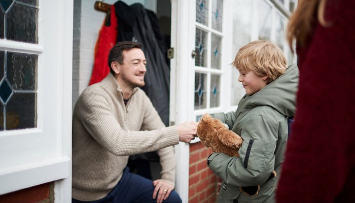 An adult kneeling at a doorway, handing an object to a child holding a teddy bear, suggesting a moment of interaction or gift exchange. The setting includes a brick wall, glass-paneled door, and coats hanging on hooks, indicating a home entrance scene.