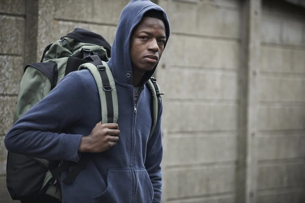 Boy with a rucksack walking outdoors, concrete wall behind him, looks at camera with serious expression