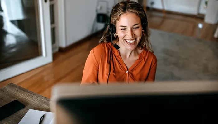 A person wearing an orange blouse and headset sitting at a desk with a computer monitor in the foreground. The setting appears to be a home office with wooden floors, a rug, and shelves in the background, suggesting remote work or online communication.