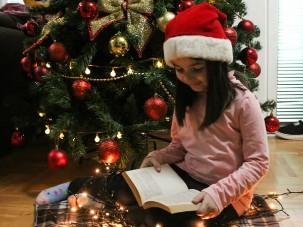 A child wearing a Santa hat and pink sweatshirt sitting on the floor near a decorated Christmas tree with red ornaments and string lights. The child is reading an open book with additional string lights scattered around, creating a cozy holiday atmosphere