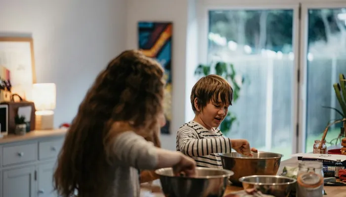 Two children standing at a kitchen table engaged in mixing ingredients in metal bowls, suggesting a baking or cooking activity. The setting includes a softly lit room with a window in the background, a dresser and various cooking items on the table