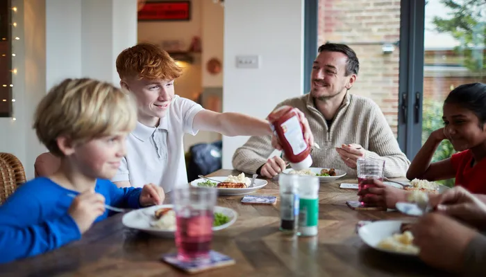 A family meal showing four people seated around a wooden table with plates of food and drinks. One person is reaching to pour ketchup, while condiments and glasses with red beverages are visible, highlighting a casual dining setting.