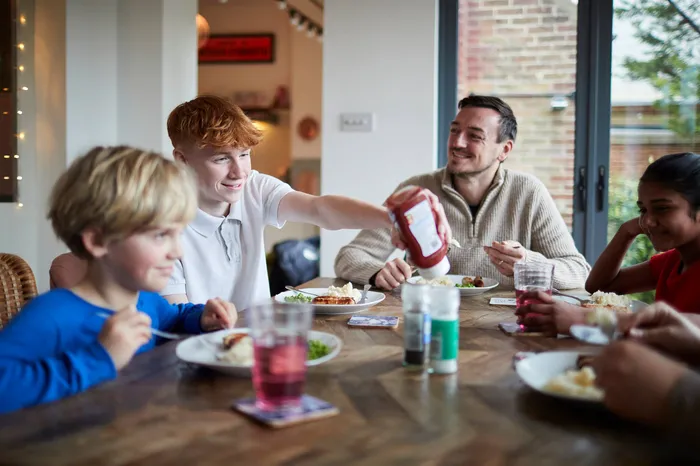 A family meal showing four people seated around a wooden table with plates of food and drinks. One person is reaching to pour ketchup, while condiments and glasses with red beverages are visible, highlighting a casual dining setting.