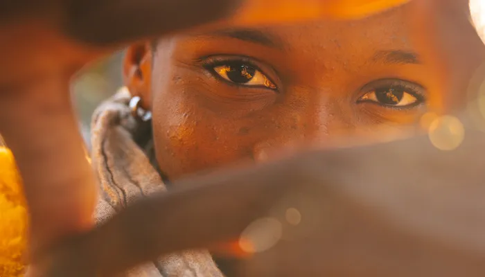 Close up showing a face of young person with their hand extended toward the camera making a frame with their fingers. The background features warm lighting and blurred elements, suggesting a sunlit atmosphere.