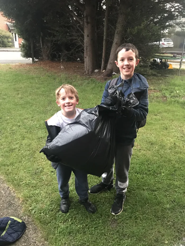Two children standing on grass outdoors, holding a large black bin bag together. Background includes trees, a road, and parked vehicles, suggesting a clean-up or outdoor activity.