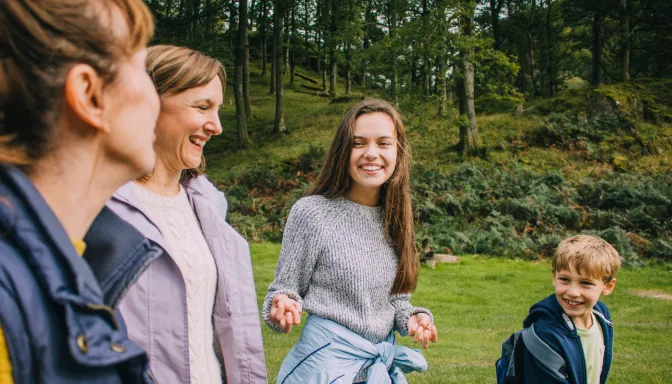 Two adults and two children outdoors in a grassy, wooded area, engaged in conversation and smiling. The individuals wear casual clothing suitable for cool weather, with trees and a hillside in the background indicating a natural, park-like setting.