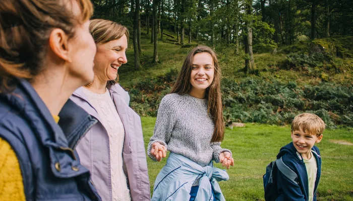 Two adults and two children outdoors in a grassy, wooded area, engaged in conversation and smiling. The individuals wear casual clothing suitable for cool weather, with trees and a hillside in the background indicating a natural, park-like setting.
