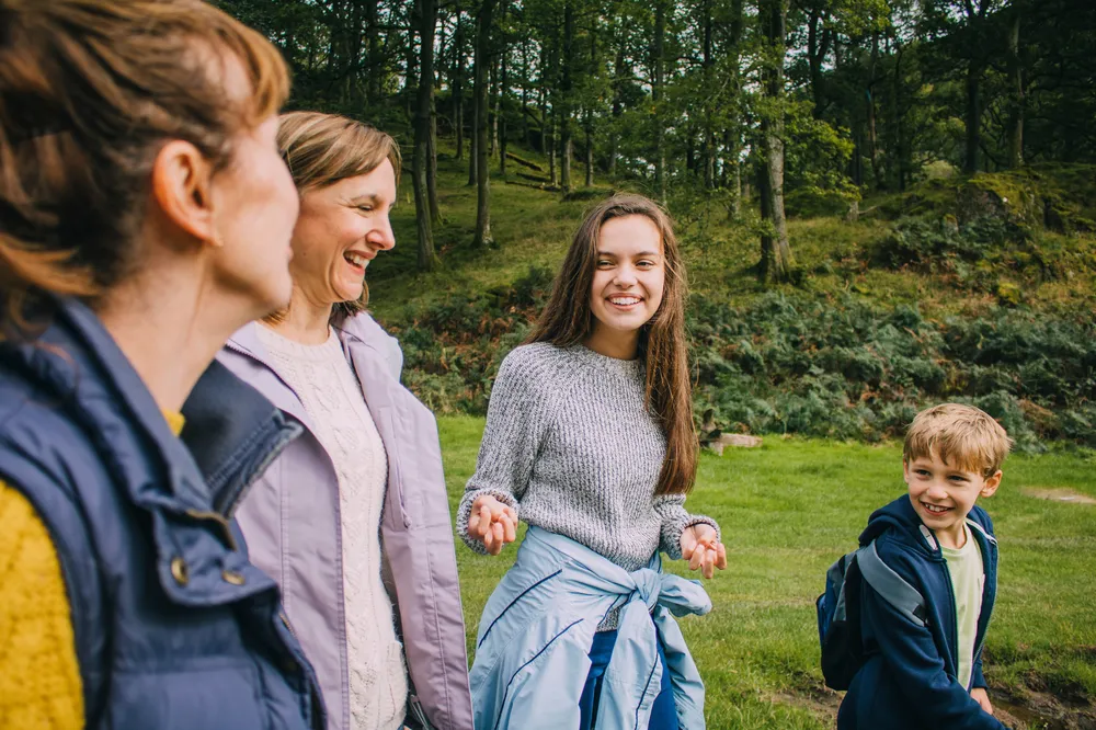 Two adults and two children outdoors in a grassy, wooded area, engaged in conversation and smiling. The individuals wear casual clothing suitable for cool weather, with trees and a hillside in the background indicating a natural, park-like setting.